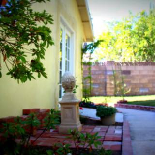 A scenic garden view featuring a pathway beside a yellow house with lush greenery and a decorative statue.