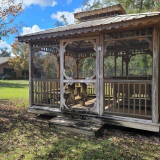 A wooden gazebo standing in a grassy area surrounded by autumn trees under a cloudy sky.
