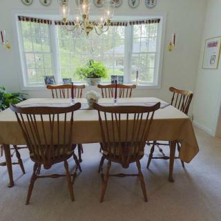 A cozy dining room with a large table, wooden chairs, and a chandelier, featuring large windows and green plants.
