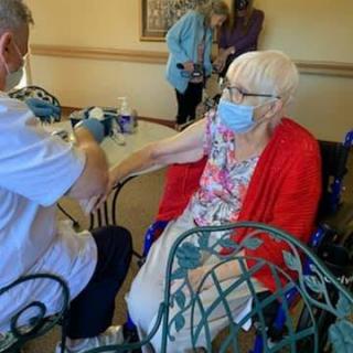 An elderly woman wearing a mask sits in a wheelchair and interacts with another person indoors.