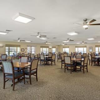 A spacious dining room with multiple tables and chairs, featuring ceiling fans and a carpeted floor.
