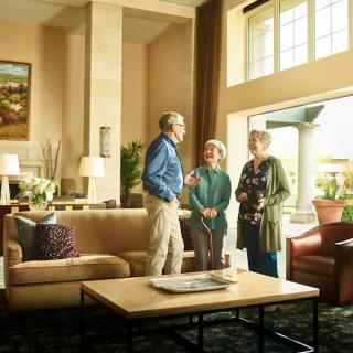 Three people engaged in conversation in a well-lit living room with large windows.