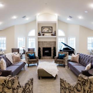 A spacious and elegantly decorated living room featuring a central fireplace, multiple sofas, armchairs, and a grand piano.