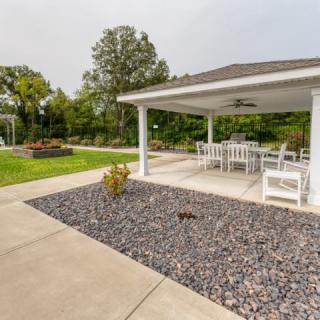 A serene outdoor patio area with white furniture and a covered gazebo, surrounded by landscaped gardens and a paved walkway.