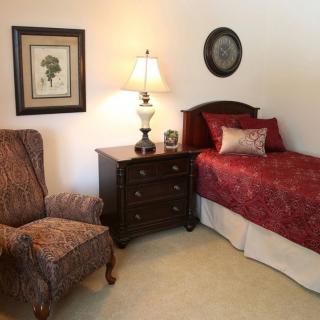 A cozy bedroom with a single bed, nightstand, lamp, and a comfortable chair.