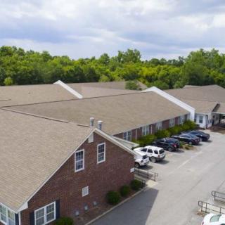 Aerial view of a brick building with a parking lot and cars, surrounded by trees under a cloudy sky.