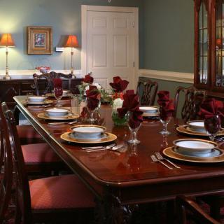 An elegant dining room featuring a long wooden table set with fine dinnerware and red floral centerpieces.