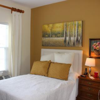 A cozy bedroom with a white bed, two yellow pillows, and wall art above the headboard. A wooden nightstand with a lamp and decorative items is beside the bed.