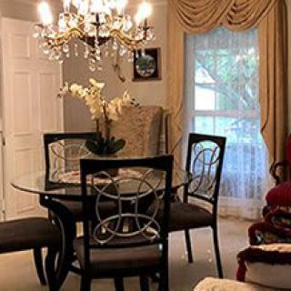 A dining room with a glass table and chairs, illuminated by an elegant chandelier, featuring a large window with draped curtains.