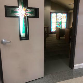 An open church door featuring a cross design, leading into a sanctuary with stained glass windows.