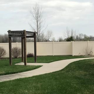 A curved pathway leads through a grassy area with a wooden archway, bordered by a tall fence.