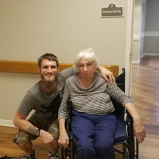A young man poses with an elderly woman in a wheelchair, showcasing a moment of care and companionship.
