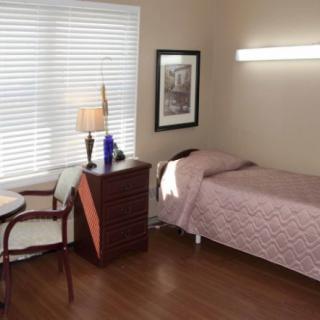 A minimalist bedroom with a single bed, bedside table, and chair, featuring natural lighting from a window.