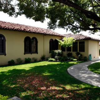 A charming house with a red-tiled roof surrounded by a lush garden and shaded by large trees.