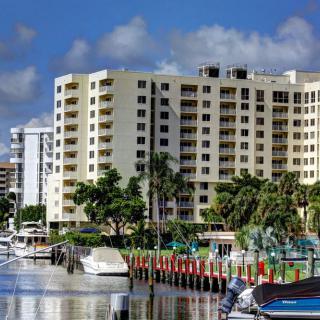 A scenic waterfront view featuring a residential apartment building with boats docked nearby and palm trees lining the shore.