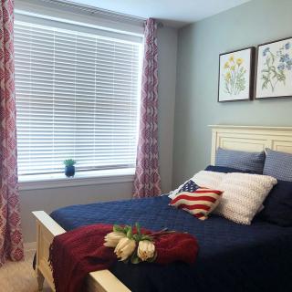 A cozy bedroom with a patriotic-themed bedspread and decor, featuring a large window with drawn curtains and elegant artwork above the bed.