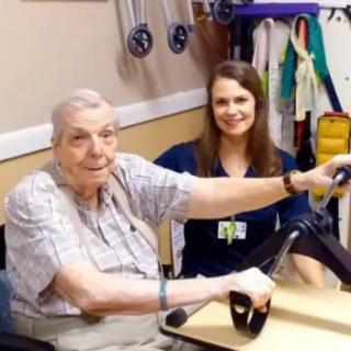 An elderly man using exercise equipment with the assistance of a healthcare professional.