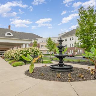A beautifully landscaped garden featuring a central fountain, surrounded by greenery and buildings under a bright blue sky.