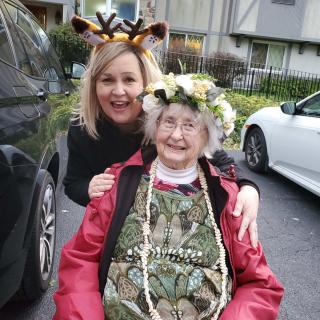 An elderly woman with a flower crown sits in a wheelchair, accompanied by a smiling younger woman wearing a giraffe headband.