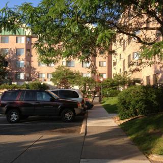 A parking lot with several cars in front of an apartment building surrounded by trees.