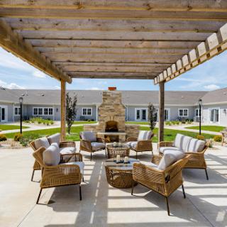 A cozy outdoor seating area featuring a stone fireplace, surrounded by comfortable chairs and a wooden pergola.