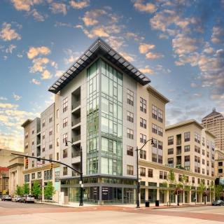 A modern urban apartment building with a glass facade, set against a backdrop of a partly cloudy sky.