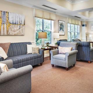 A cozy living room with comfortable sofas and armchairs, featuring a carpeted floor and large windows accented by elegant decor.