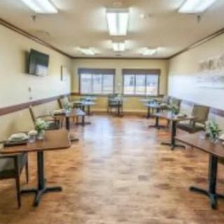 A clean and well-lit dining room with several tables and chairs, featuring large windows and a wooden floor.