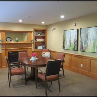 A cozy conference room with a round table surrounded by chairs, featuring artwork on the walls and a bookshelf in the corner.