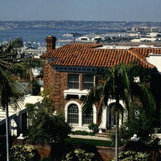 A beautiful Mediterranean-style house with a red-tiled roof, surrounded by palm trees and overlooking a scenic sea view.