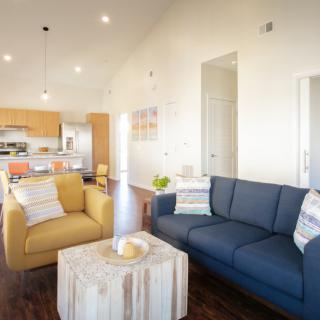 A modern living room with bright lighting, featuring a blue and yellow sofa, wooden floors, and an open kitchen layout.