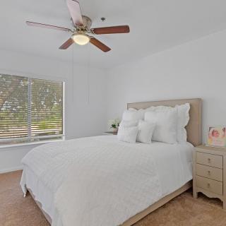 A cozy bedroom with a neatly made bed, decorated with white bedding and pillows, featuring a ceiling fan and a window allowing natural light.
