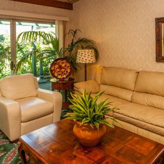 A cozy living room featuring a beige sofa, armchair, wooden coffee table with a potted plant, and a large window with outdoor greenery.