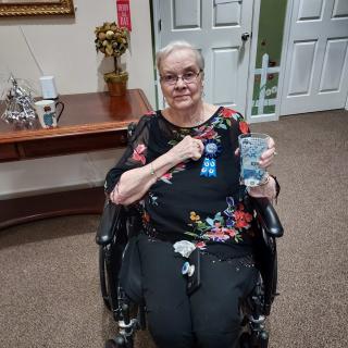 An elderly woman seated in a wheelchair displays an award ribbon while smiling indoors.