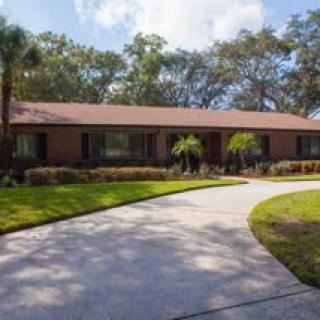 A suburban single-story house with a curved driveway and well-maintained lawn surrounded by trees.