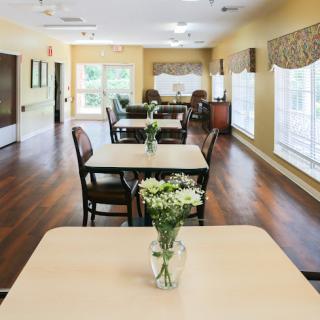 A welcoming dining area with tables set for guests, featuring natural light and a floral centerpiece.