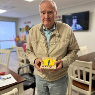 An elderly man holding a decorated yellow cake in an indoor setting.