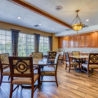 A cozy dining room with wooden floors, elegant tables and chairs, ample natural light from large windows, and a decorative chandelier.