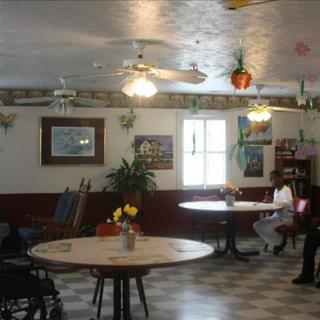 An interior view of a restaurant with round tables, chairs, and decor, including plants and paintings. A person is seated at one of the tables.