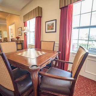 A cozy dining room with a wooden table and upholstered chairs, featuring large windows with red curtains.