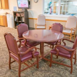 A waiting room with a round table surrounded by chairs, a television on the wall, and a carpeted floor.