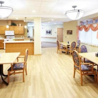 A bright dining area with tables and chairs, featuring a view of the kitchen and wooden flooring.