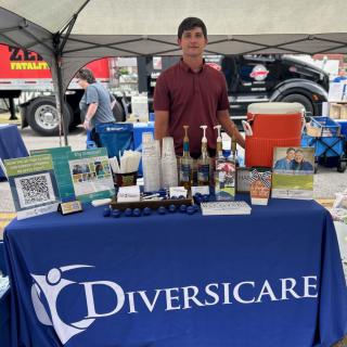 A person stands behind a Diversicare booth at an outdoor event, showcasing various informational materials and coffee supplies.