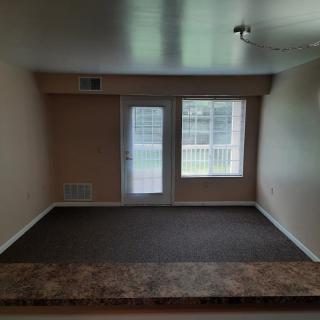 An empty living room with carpet flooring, featuring large windows and doors allowing natural light.