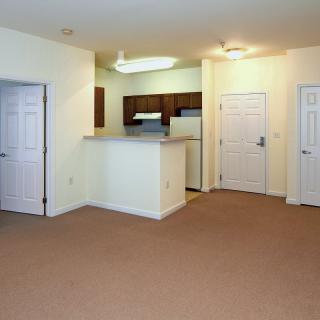 An unfurnished apartment with a small kitchen area, white walls, and carpeted flooring.