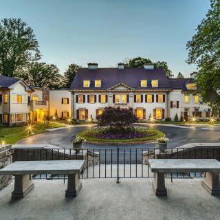 A grand mansion with illuminated exterior and central fountain, surrounded by trees during the evening.