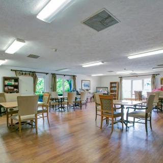 A spacious dining area with multiple tables and chairs, featuring wood flooring and bright natural lighting from large windows.