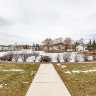 A snowy suburban landscape with a sidewalk leading to a pond, surrounded by houses and bare trees under an overcast sky.