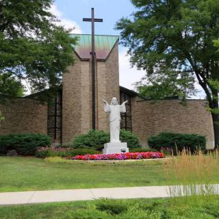 A church with a prominent cross, surrounded by greenery and a statue in the foreground.