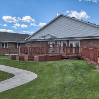 A residential building with a wooden deck, surrounded by a grassy lawn and a winding pathway under a partly cloudy sky.